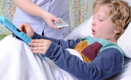 A small boy lies in a hospital bed, while a nurse holds a mobile phone near him to record his cough.